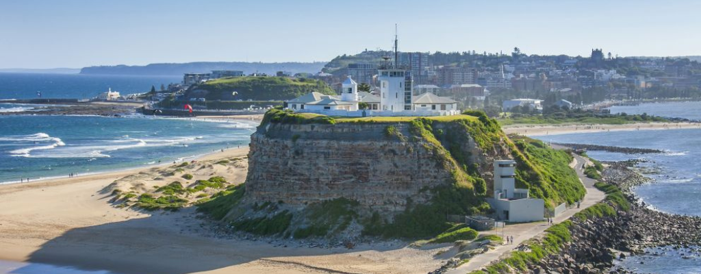 A lighthouse and lighthouse keeper cottages on top of headland surrounded by water. Newcastle city can be seen in the distance.